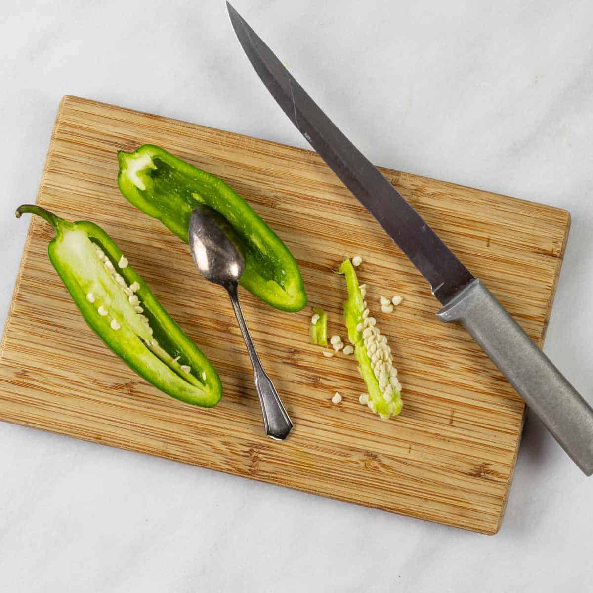 Jalapeño cut in half on a small board with a knife, spoon, and seeds partly removed.