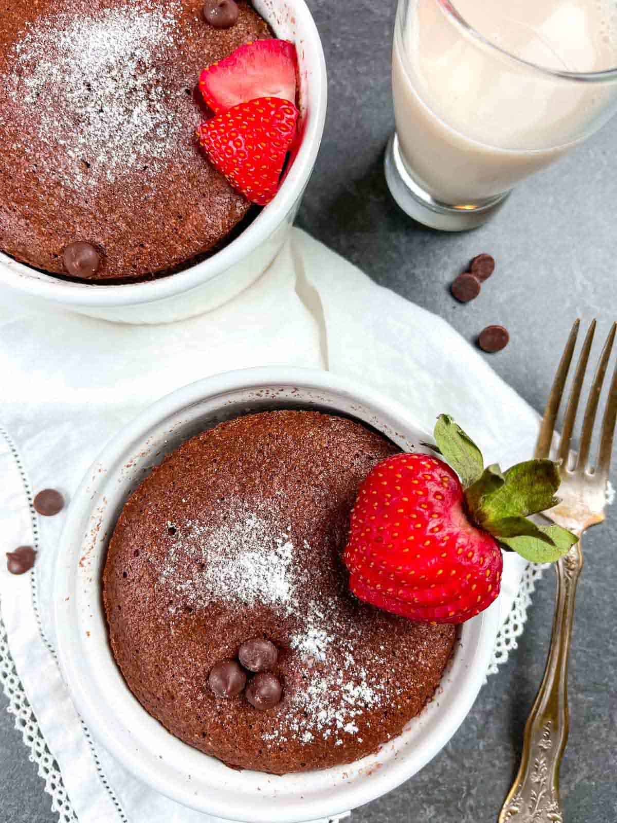 Two mug cakes in white ramekins with sliced strawberries and chocolate chips with a fork and a glass of milk.
