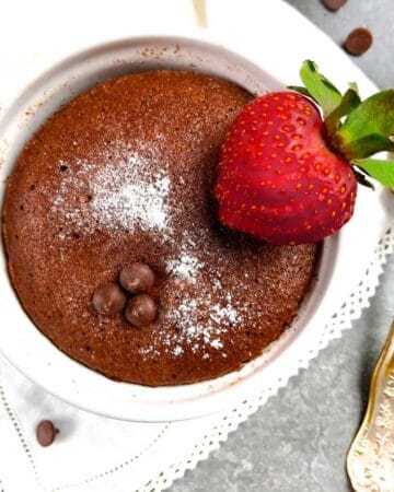 Chocolate mug cake in a white ramekin with a sliced strawberry on a white lace napkin with a gold fork.