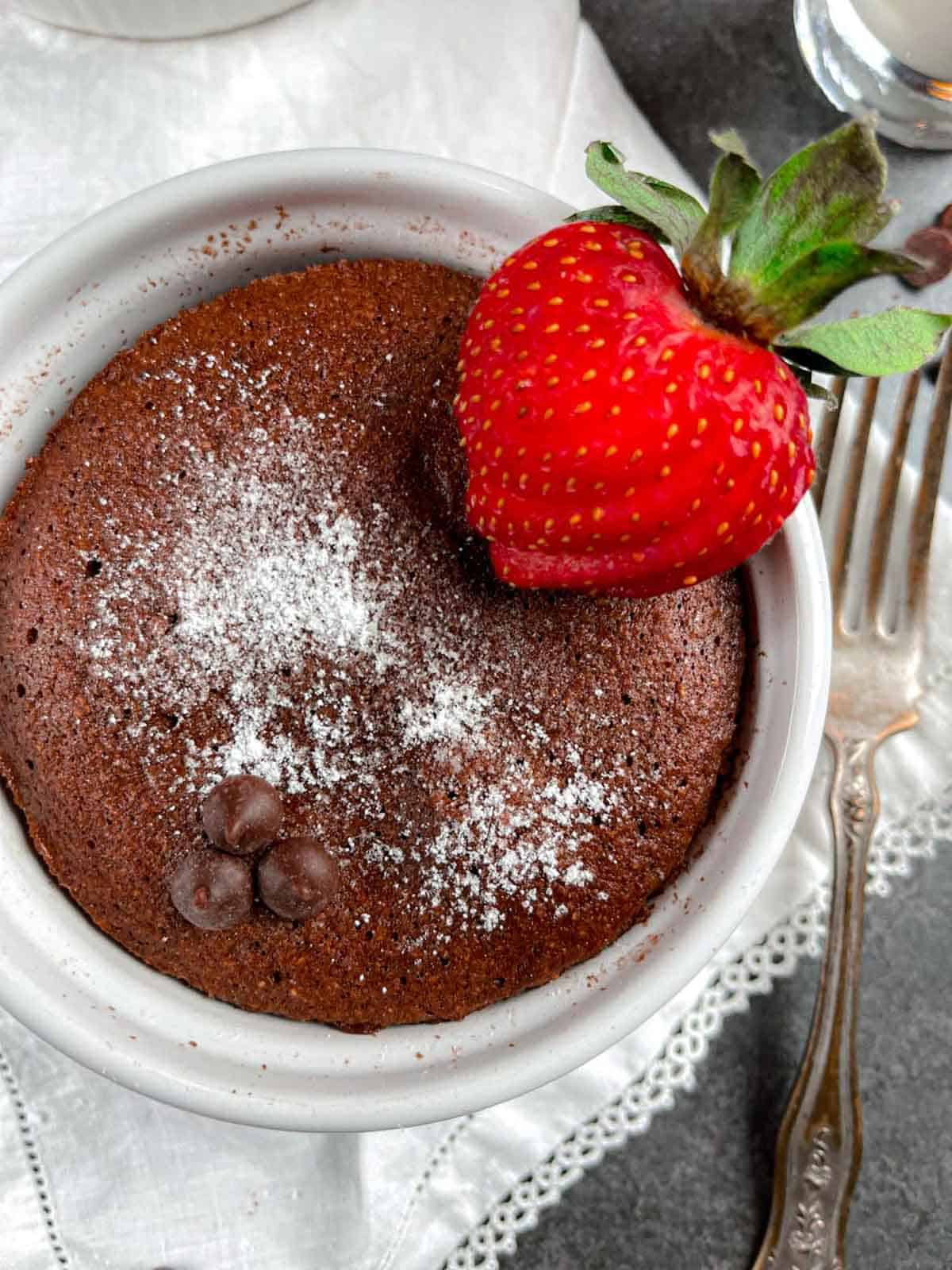 Chocolate mug cake in a white ramekin with a sliced strawberry and chocolate chips on a white lace napkin.