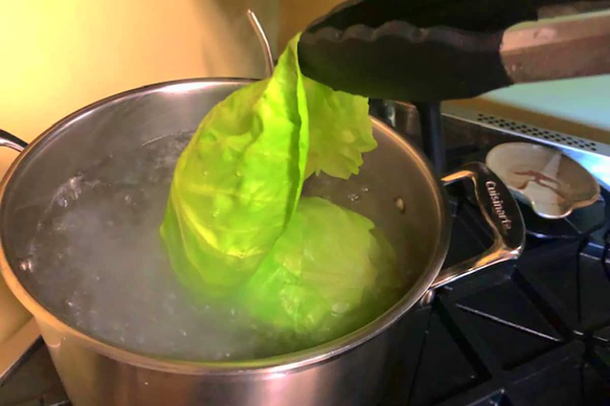 Head of cabbage in boiling water with leaves being pulled off with tongs.