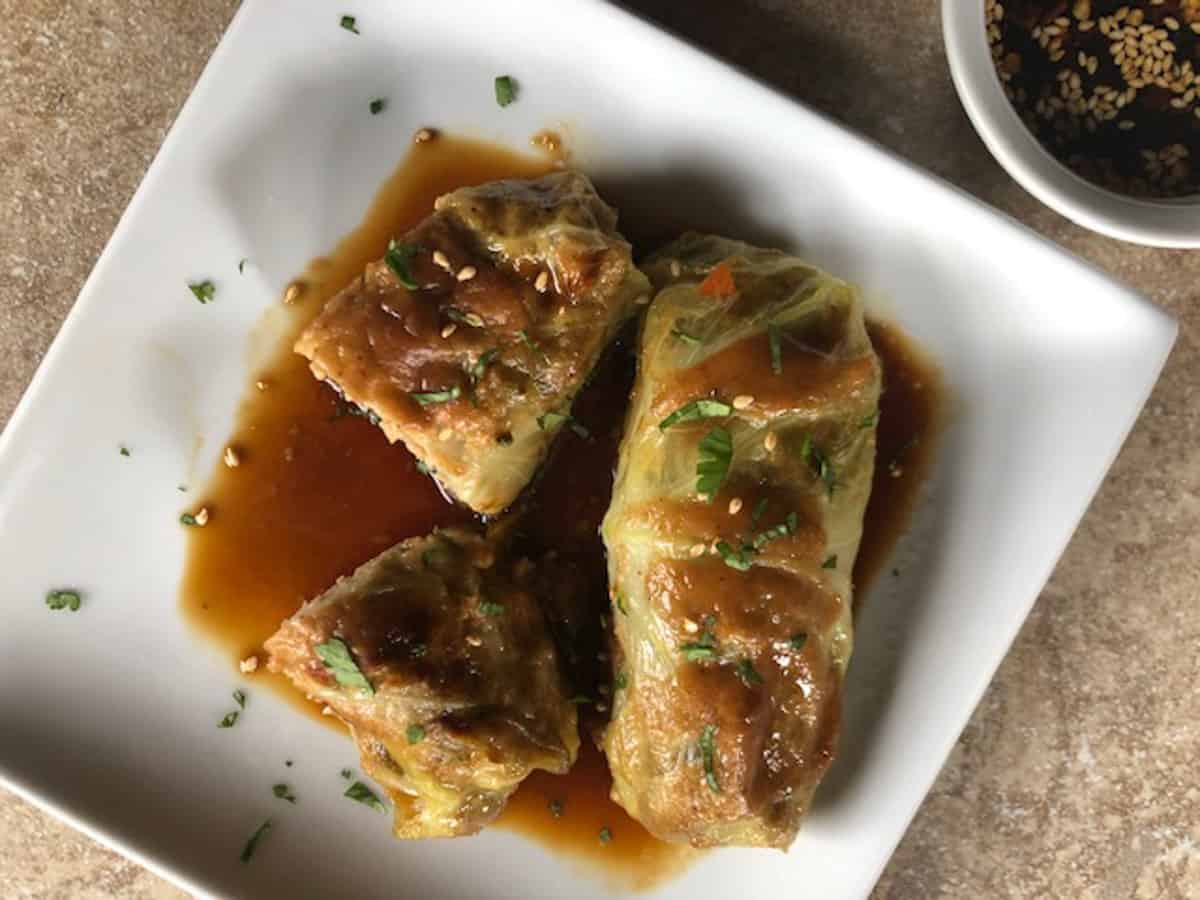 Overhead view of two baked cabbage rolls on a white square plate in a soy sauce, with one roll cut in half.