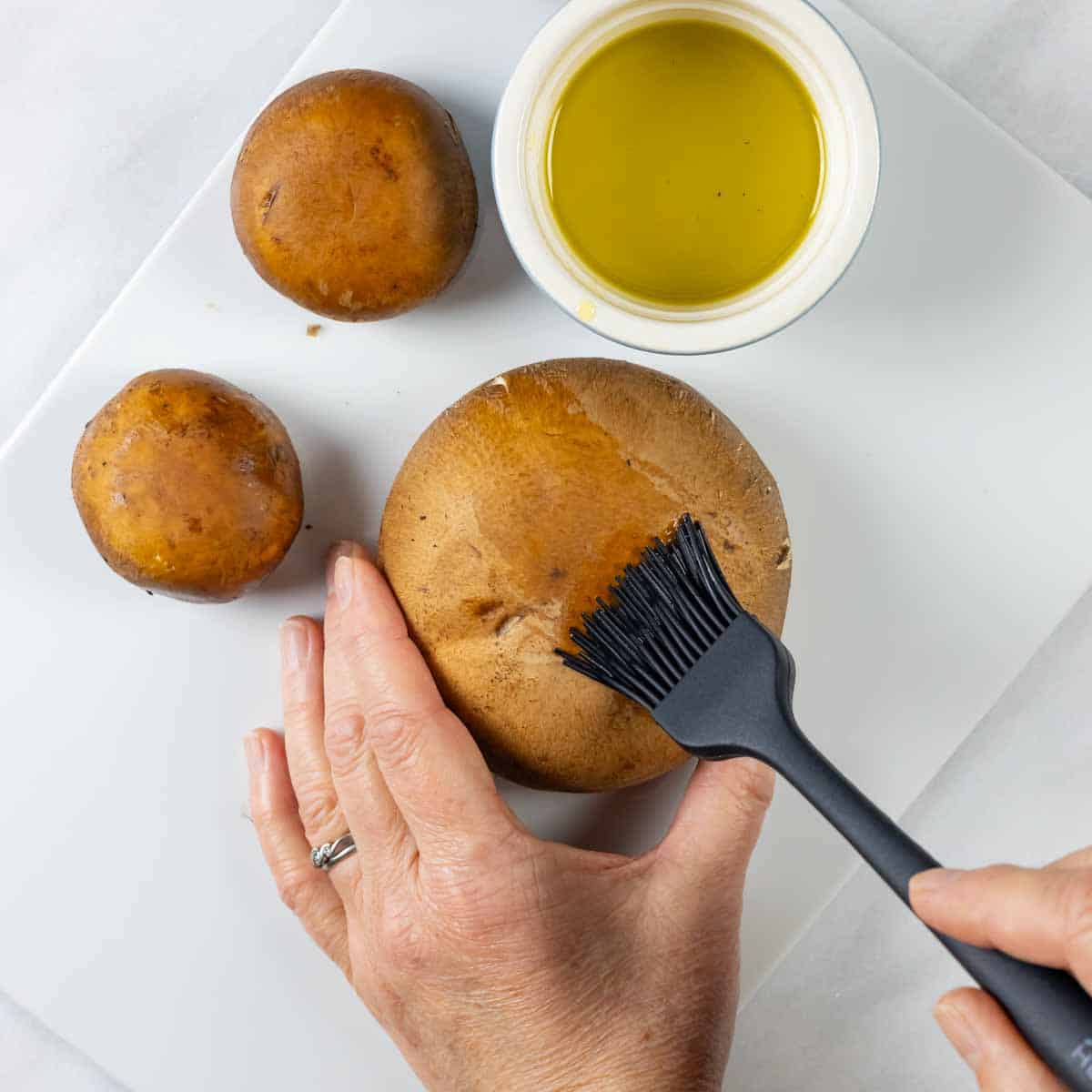Hands brushing tops of mushrooms with olive oil using a basting brush.