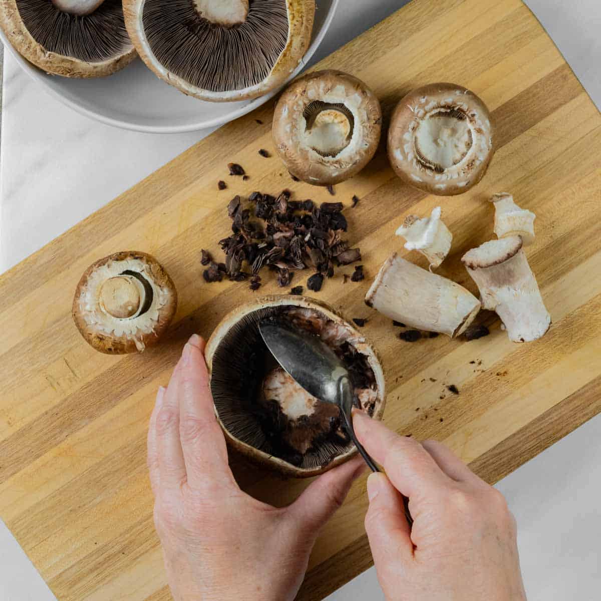 Several mushrooms on a board with stems removed and person scraping out gills with a spoon.