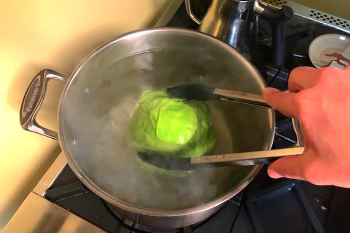 Head of cabbage boiling in a pot of water, held down in the water by a hand holding tongs.