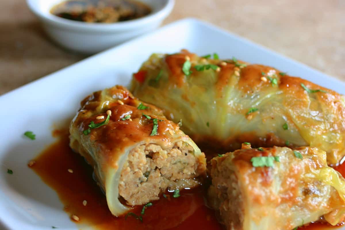 Two finished cabbage rolls on a plate covered in sauce, sesame seeds and a little parsley.  One is cut in half to show the inside.