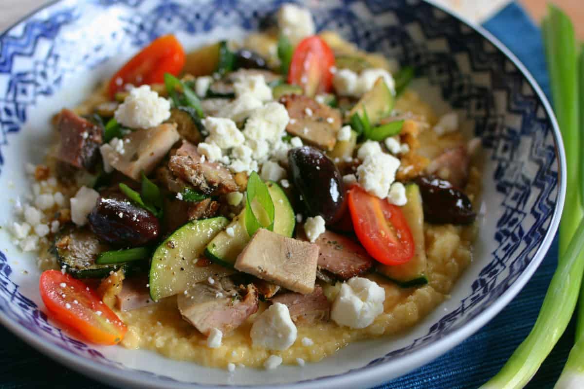 Mediterranean stir fry on a bed of keto skordalia in a white bowl with dark blue edging topped with feta cheese.