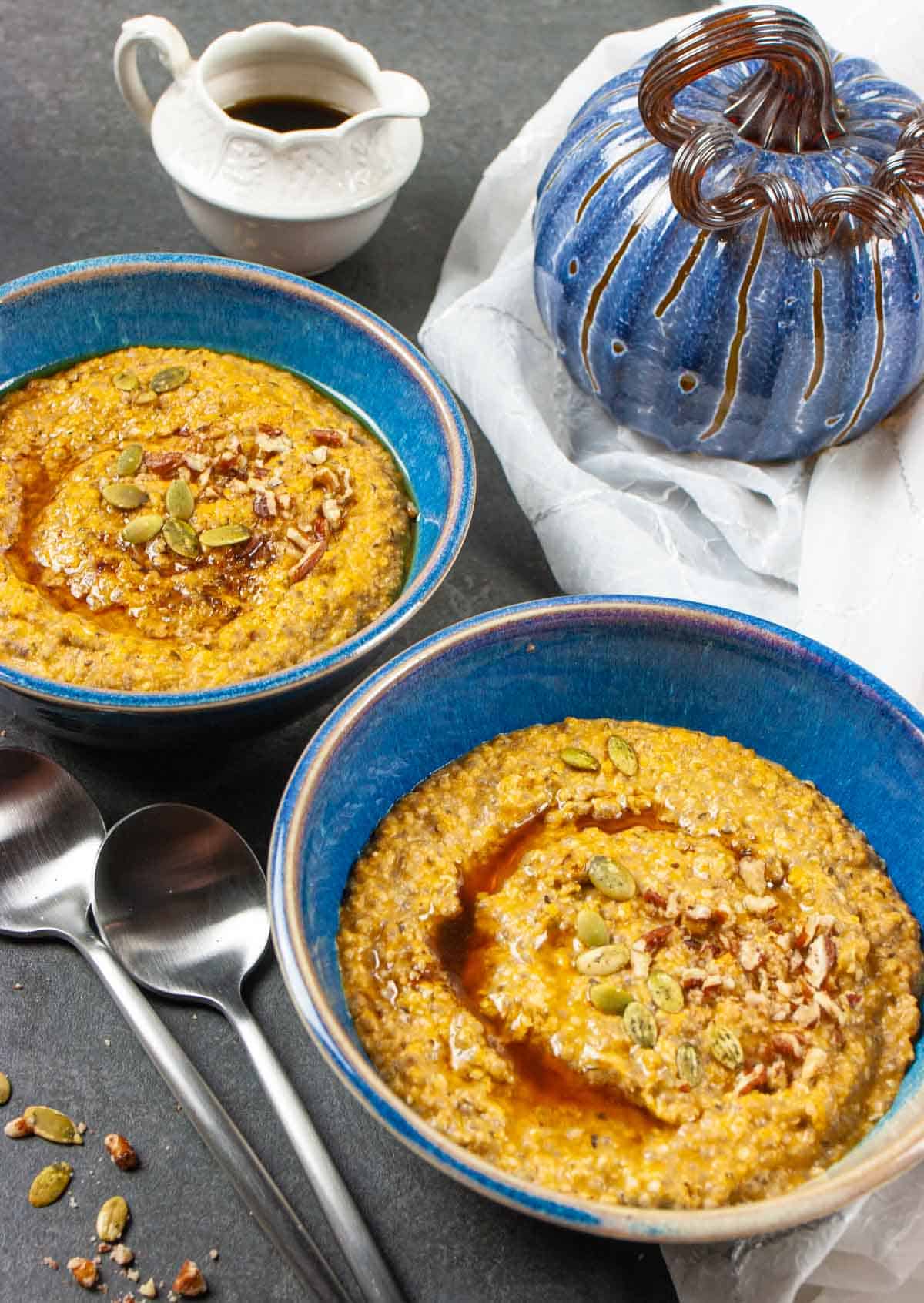 Two bowls of porridge topped with syrup, pumpkin seeds, and pecans with decorative white cup of syrup on the side and a blue blown glass pumpkin.