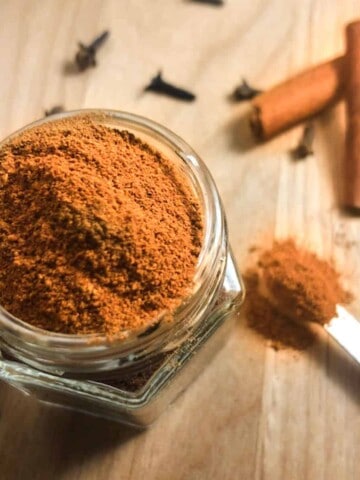 Glass spice jar with pumpkin spice seasoning, a measuring spoon full of spice, and some whole cloves and cinnamon sticks as garnish.