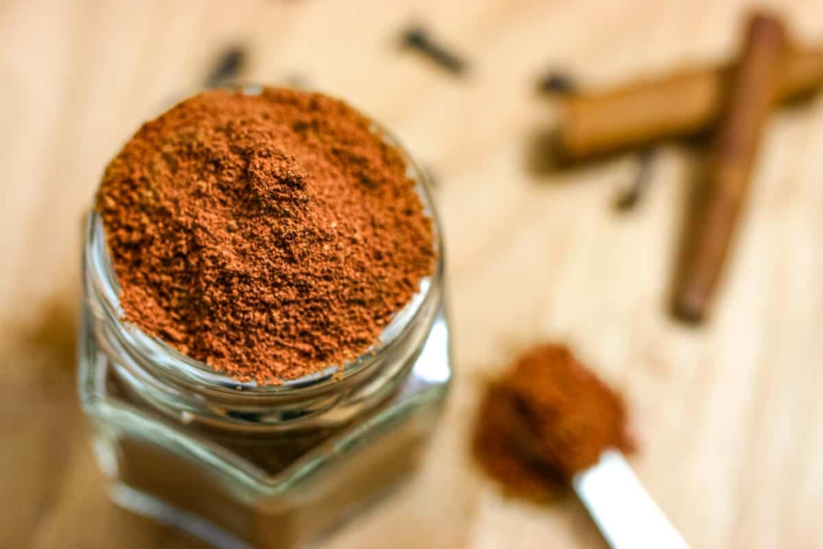 Spice jar overflowing with spice seasoning on a counter with cinnamon sticks and teaspoon of spice.