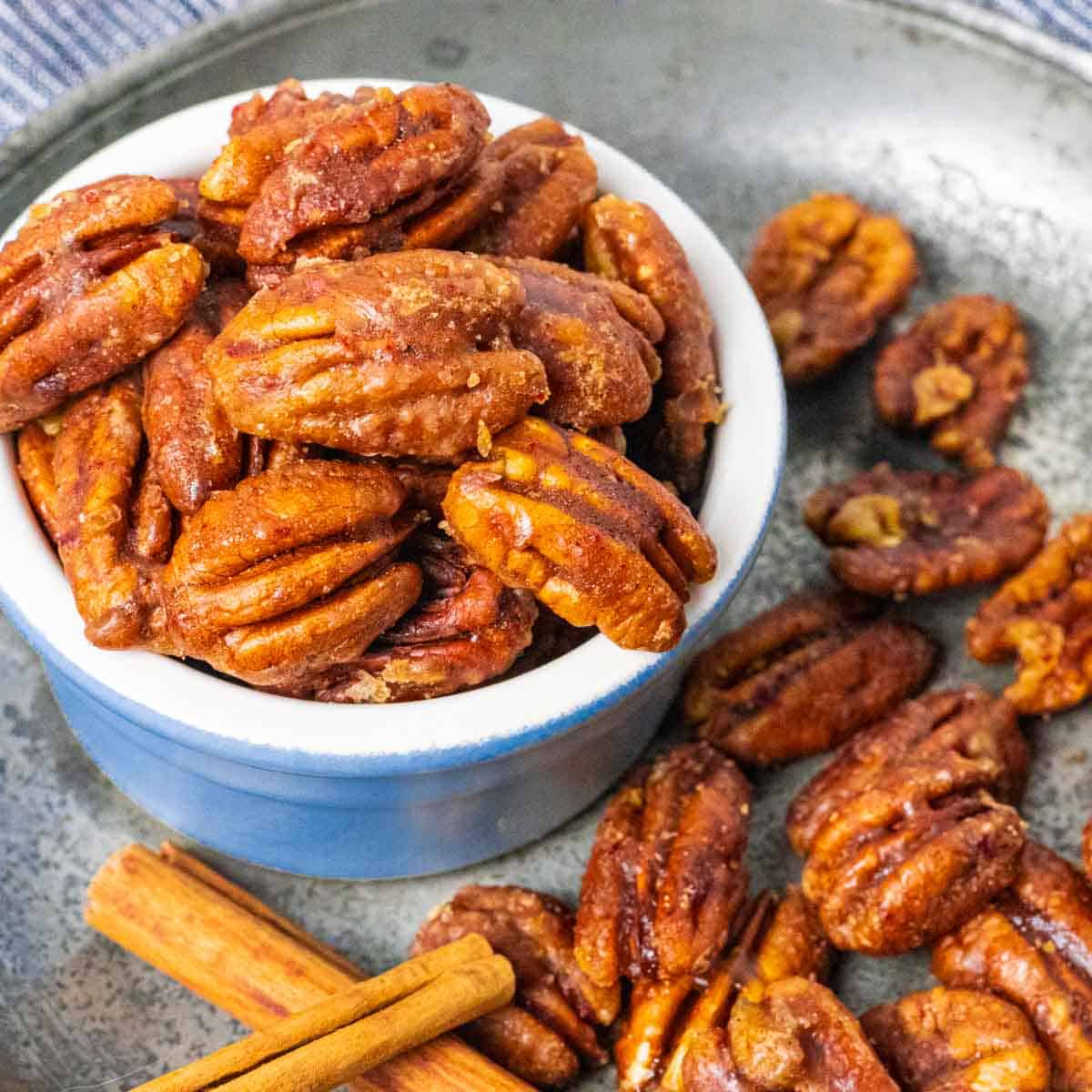Small blue and white ramekin filled with candied pecans on a board with more pecans and cinnamon sticks scattered around. 