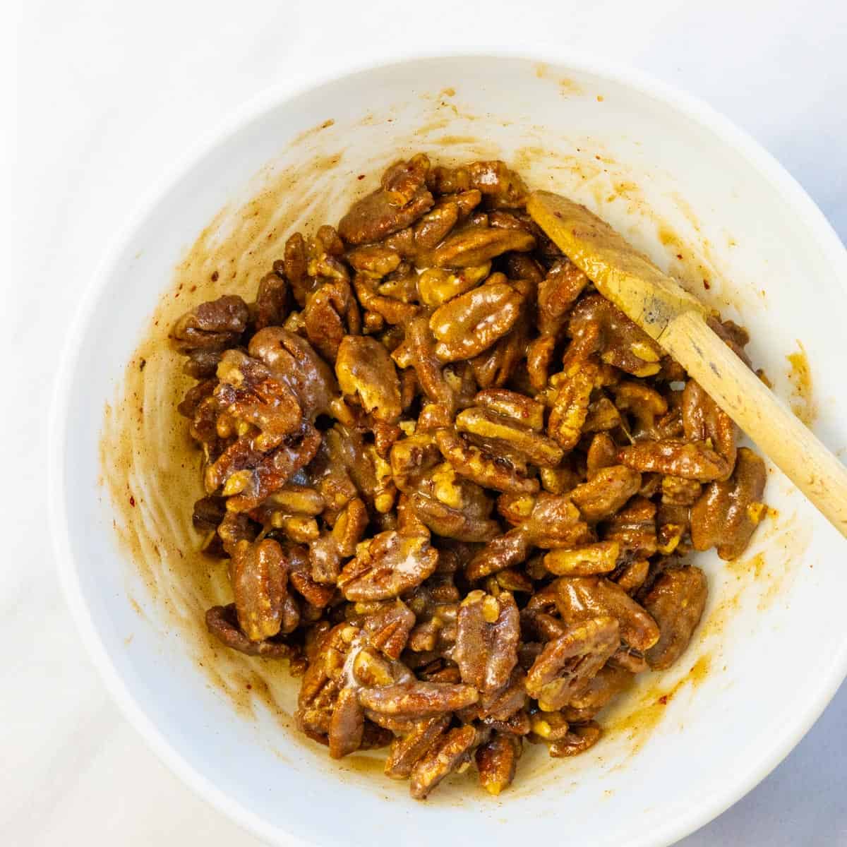 Raw pecans stirred into a mixing bowl with a wooden spoon to coat with egg white and spices.