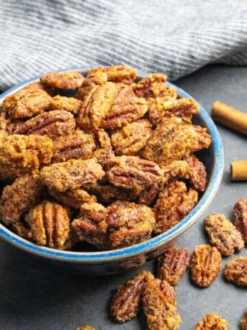 Blue bowl of bourbon candied pecans on a grey board with a towel and extra pecans and cinnamon sticks.