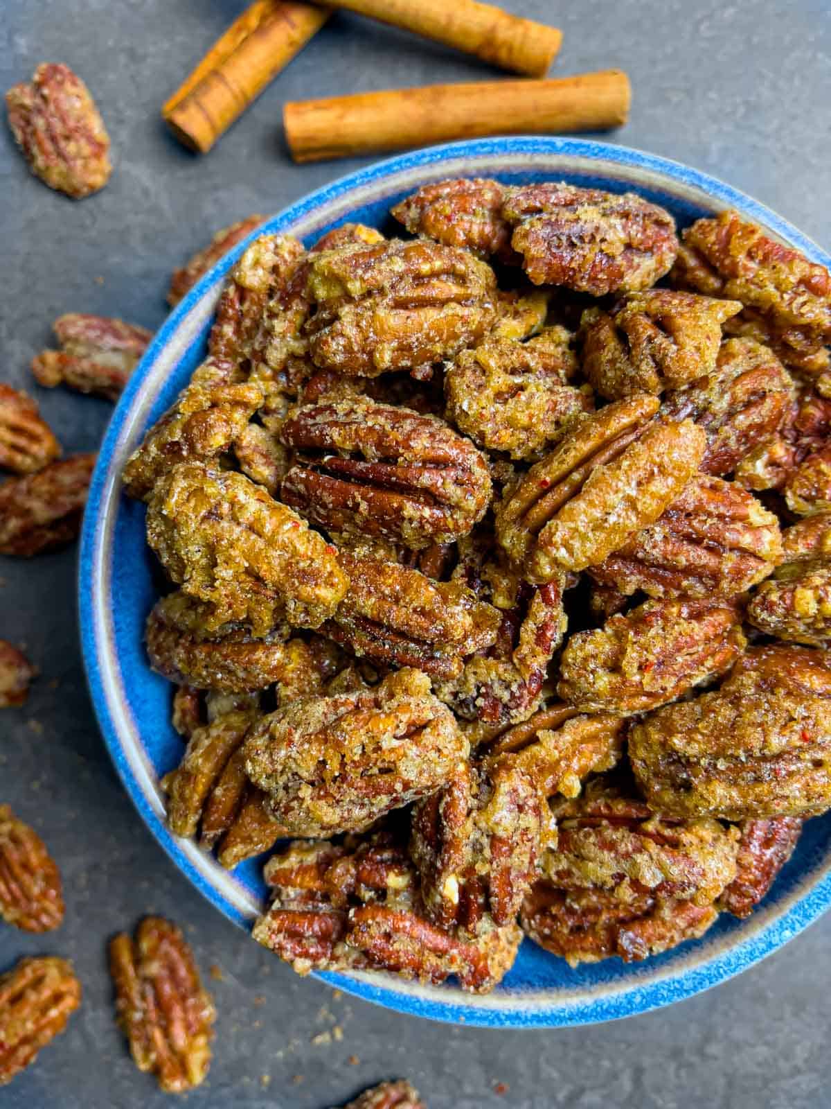 Glaze encrusted candied pecans overflowing a blue bowl with cinnamon sticks and more pecans scattered around.
