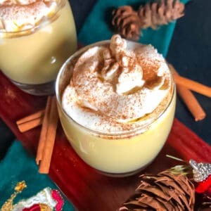 Two glasses of keto eggnog topped with cinnamon sprinkled whipped cream on a board with cinnamon sticks as garnish.