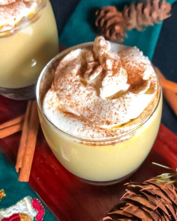 Two glasses of keto eggnog topped with cinnamon sprinkled whipped cream on a board with cinnamon sticks as garnish.