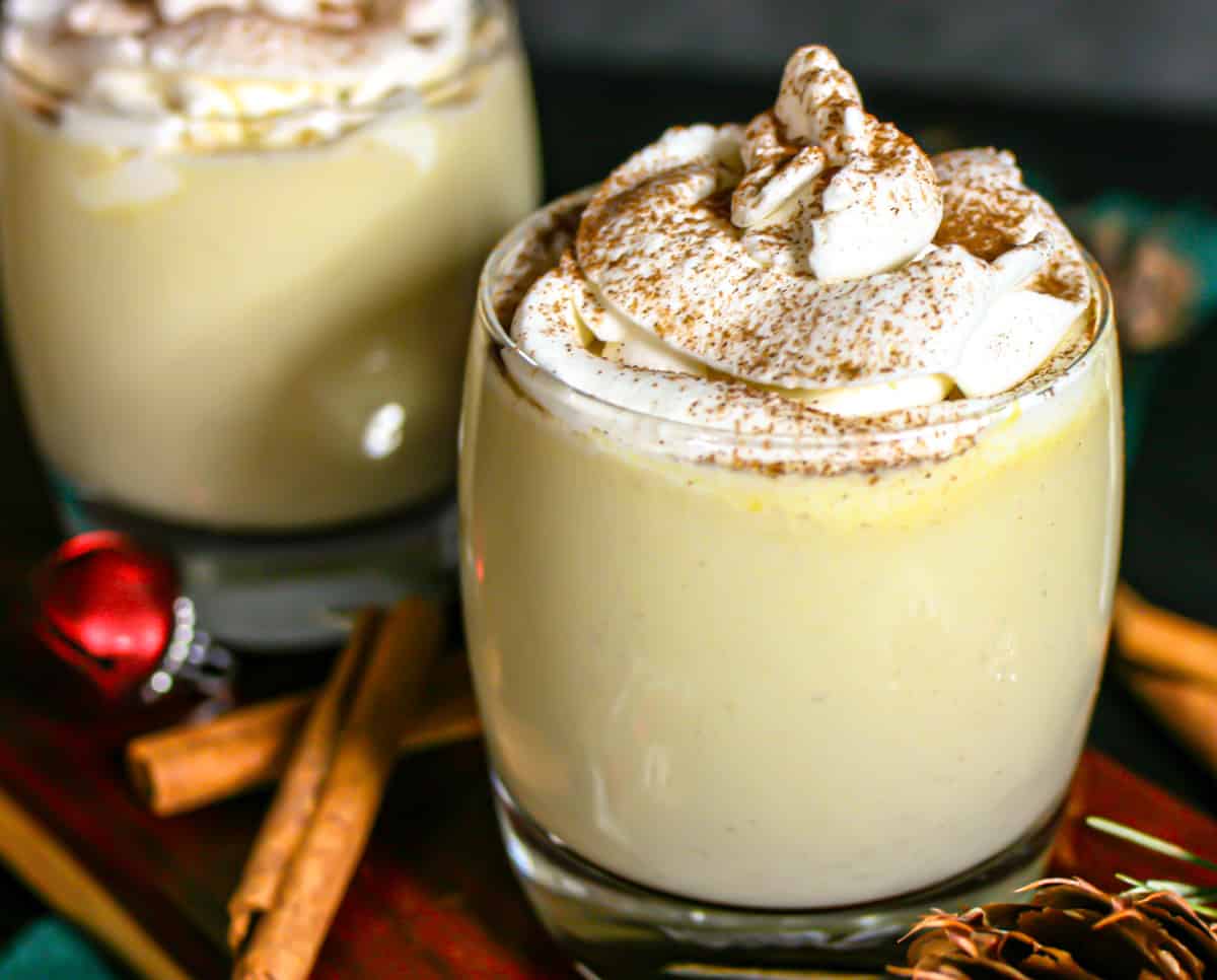 Festive glasses of holiday keto eggnog with whipped cream and cinnamon on a board with cinnamon sticks and holiday bells.