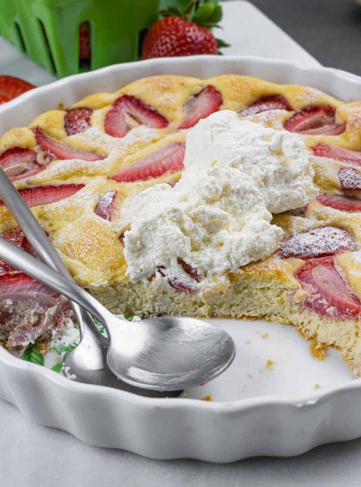 Berry clafoutis in tart dish topped with sugar-free whipped cream showing a serving removed and two spoons in the empty space.