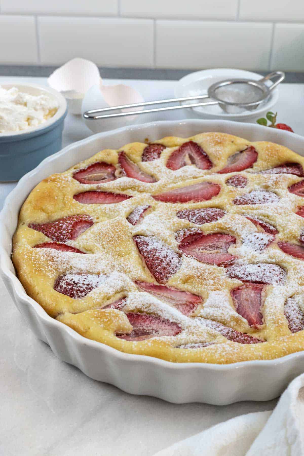 Baked clafoutis with strawberries in tart dish sprinkled with keto powdered sweetener and egg shell halves on the side.