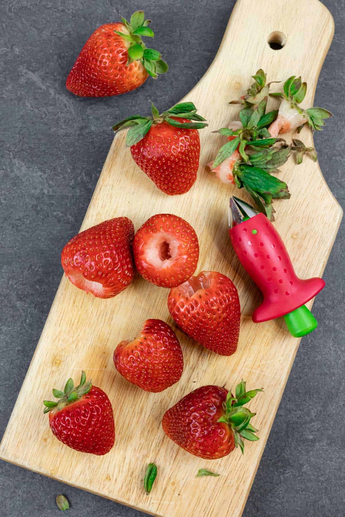 Whole and hulled strawberries and tops, on a wood board with a strawberry huller tool.