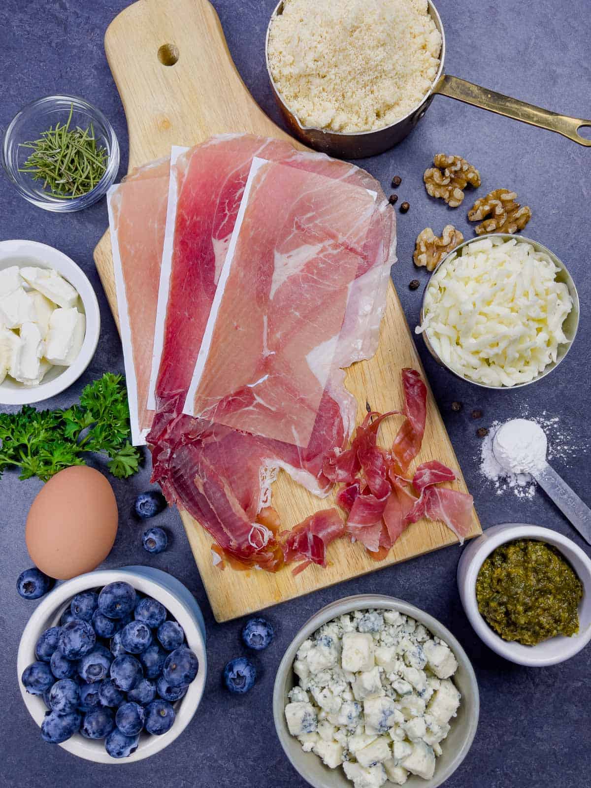 Pizza ingredients on a board including prosciutto, gorgonzola, rosemary, and blueberries in ramekins.