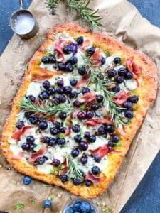 Baked blueberry pizza on parchment with sprigs of fresh rosemary on top.