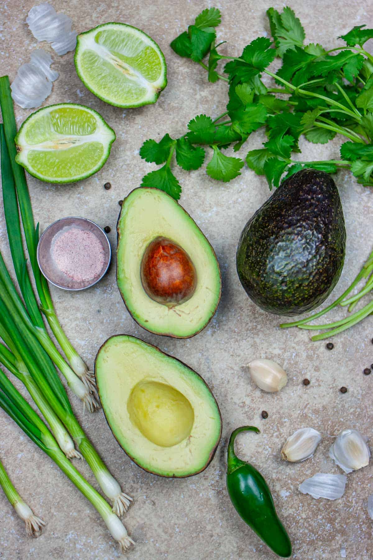Ingredients for guacamole, including avocado halves, green onions, jalapeno, garlic, cilantro, and black salt on a grey board.
