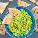 Guacamole in a blue bowl with chips in it and scattered around the bowl.