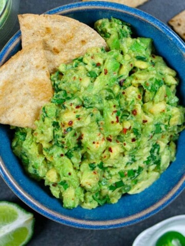 Guacamole sprinkled with Aleppo pepper in a blue bowl with a side of chips and a quarter of a lime.