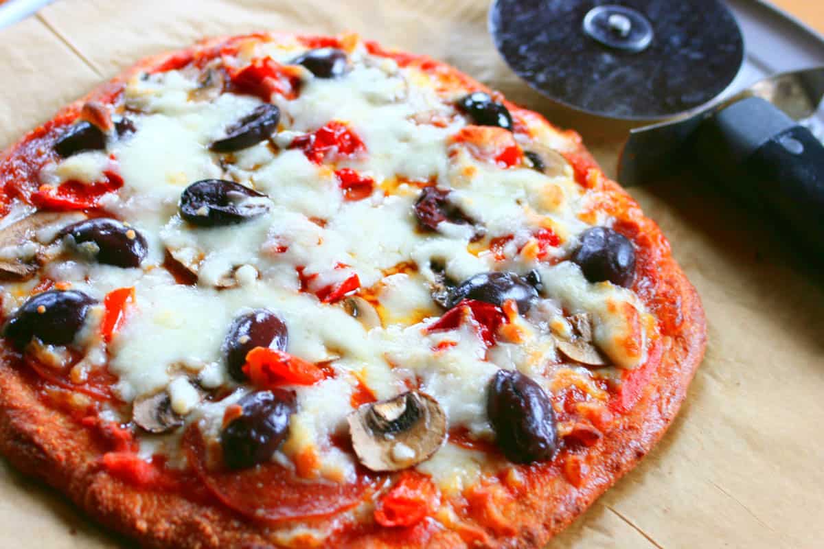 Low carb pizza crust with pepperoni, olives and mushrooms on parchment with a pizza cutter.