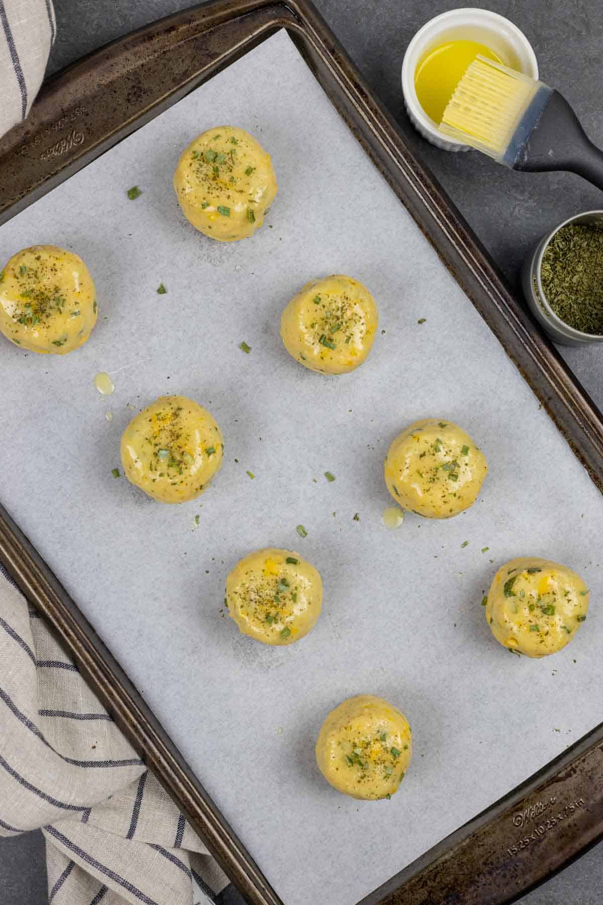 Biscuit dough buttered and sprinkled with herbs on a baking sheet ready to go in the oven.  Captioned with instructions.