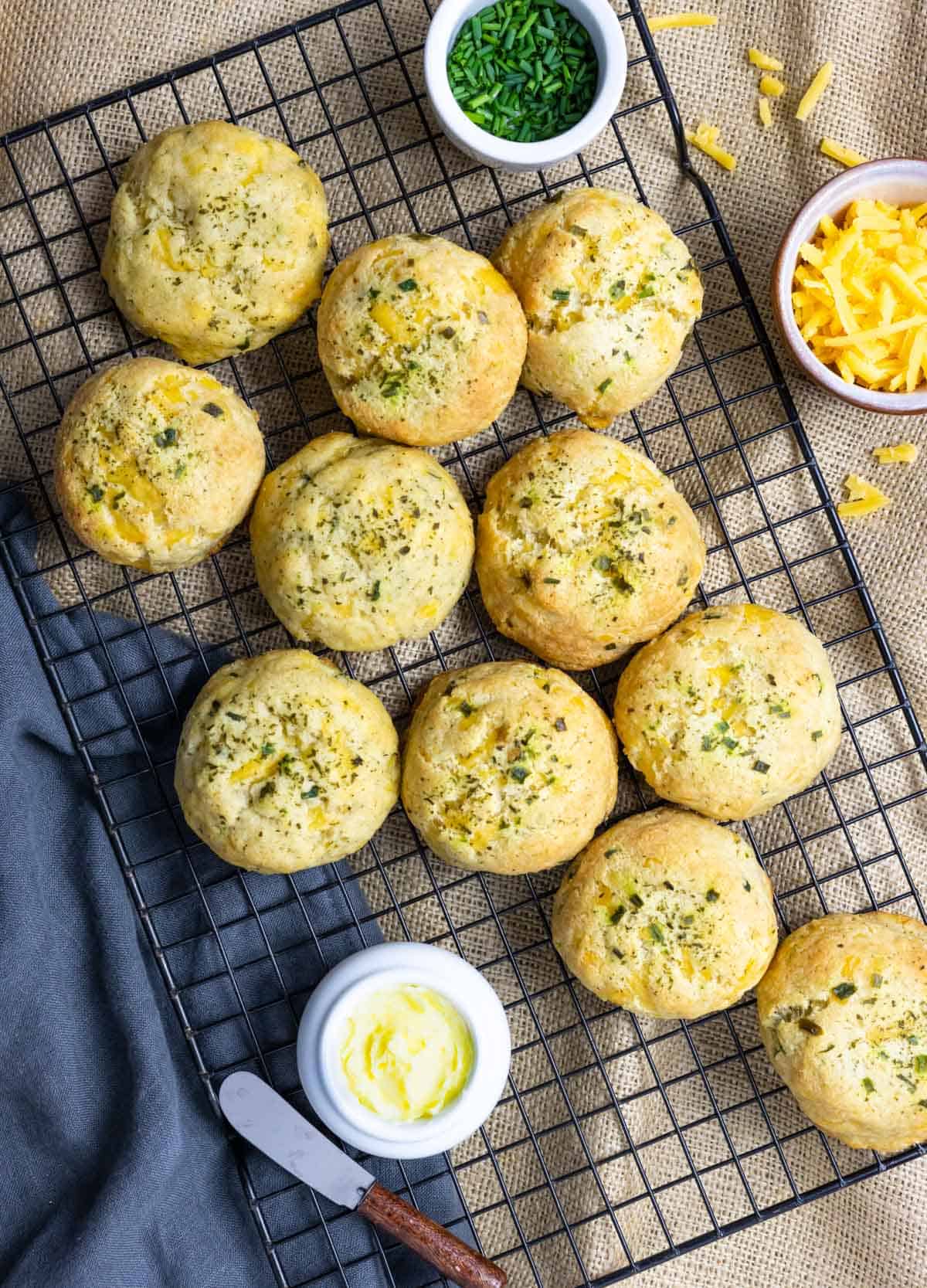 Cheddar biscuits cooling on a black rack with small cups of chives, cheese and butter on the side.