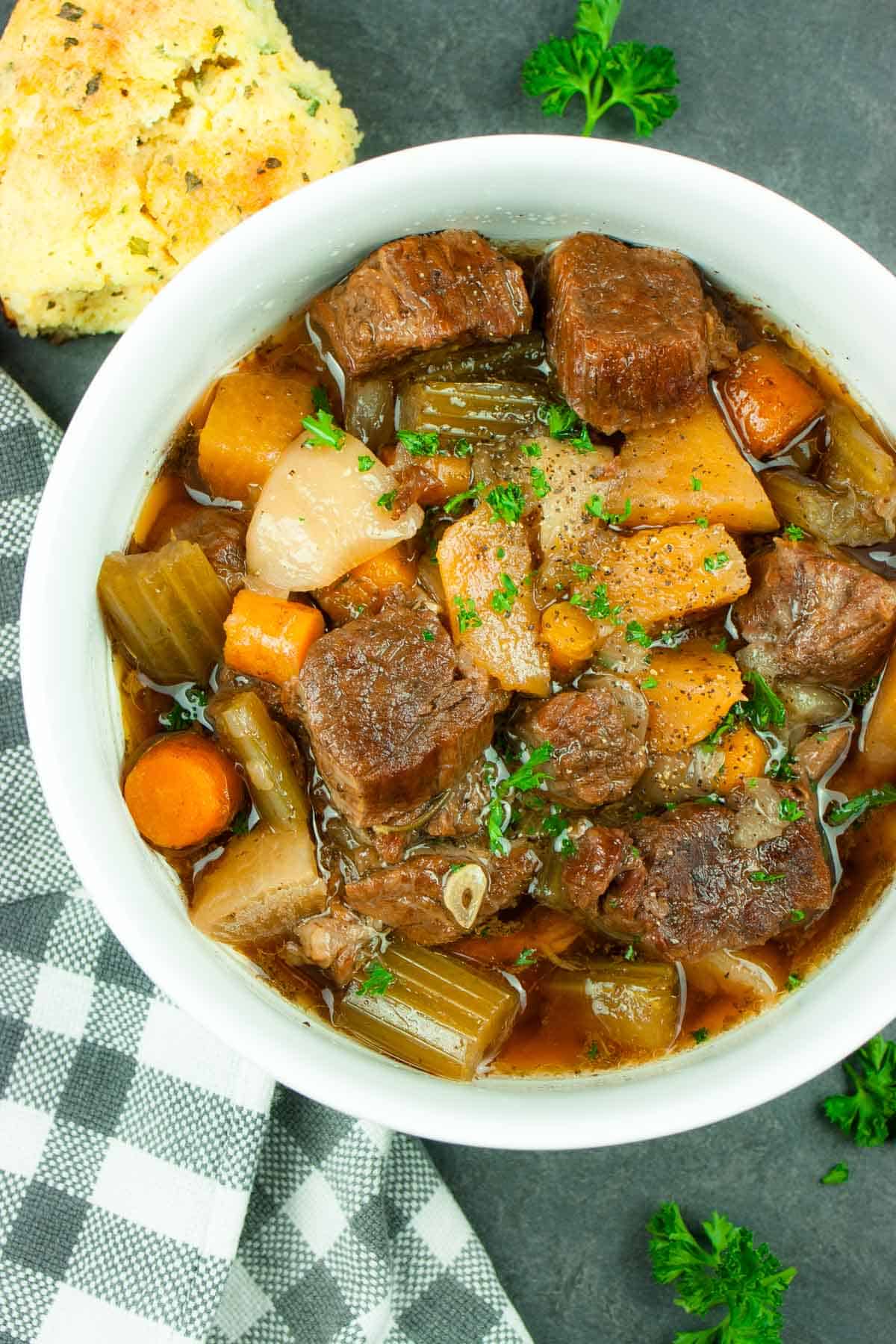 Irish beef stew in a white bowl with a spoon with a dish towel and a biscuit.