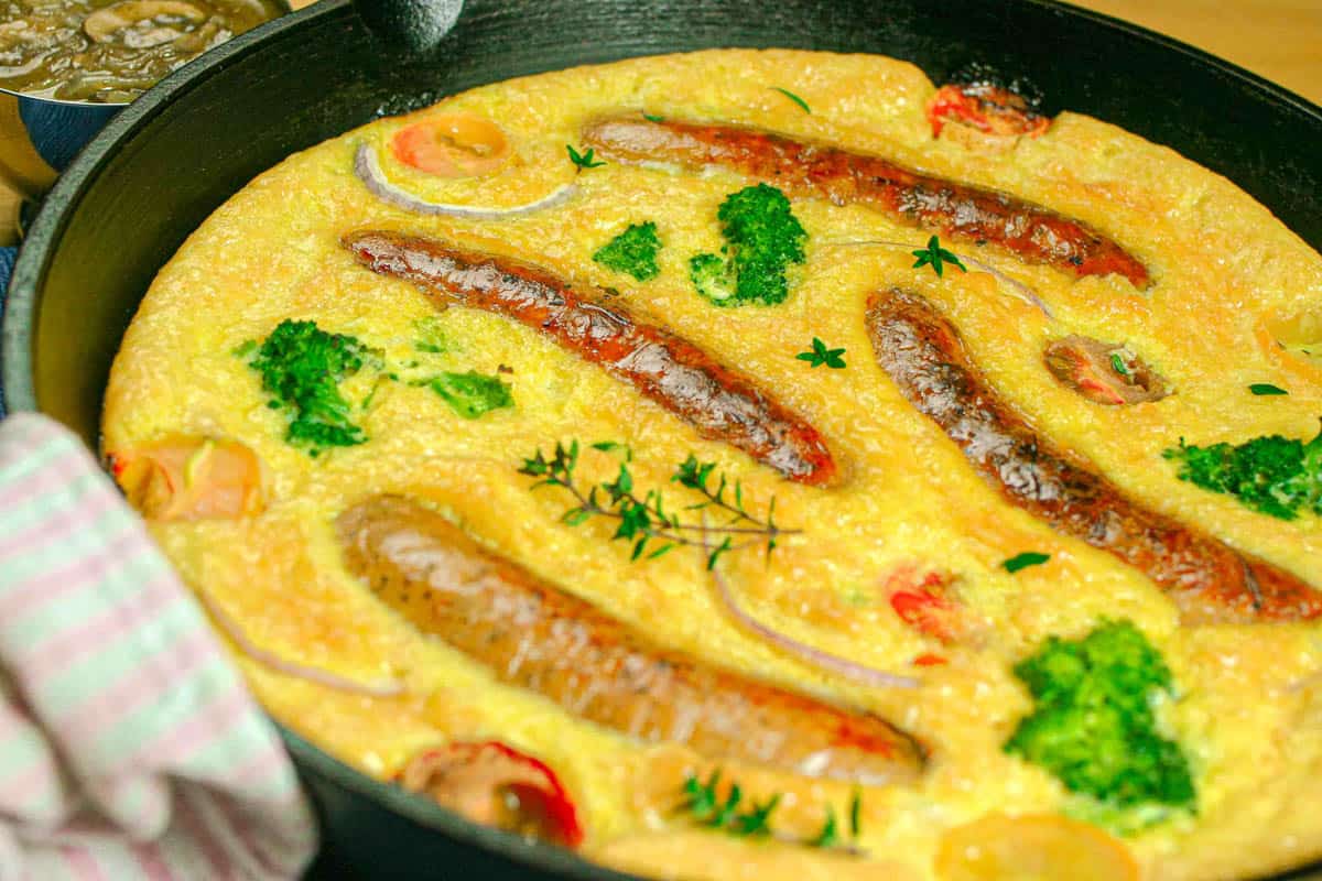 Hot cast iron skillet of baked toad in the hole with veggies and a towel wrapping the handle.