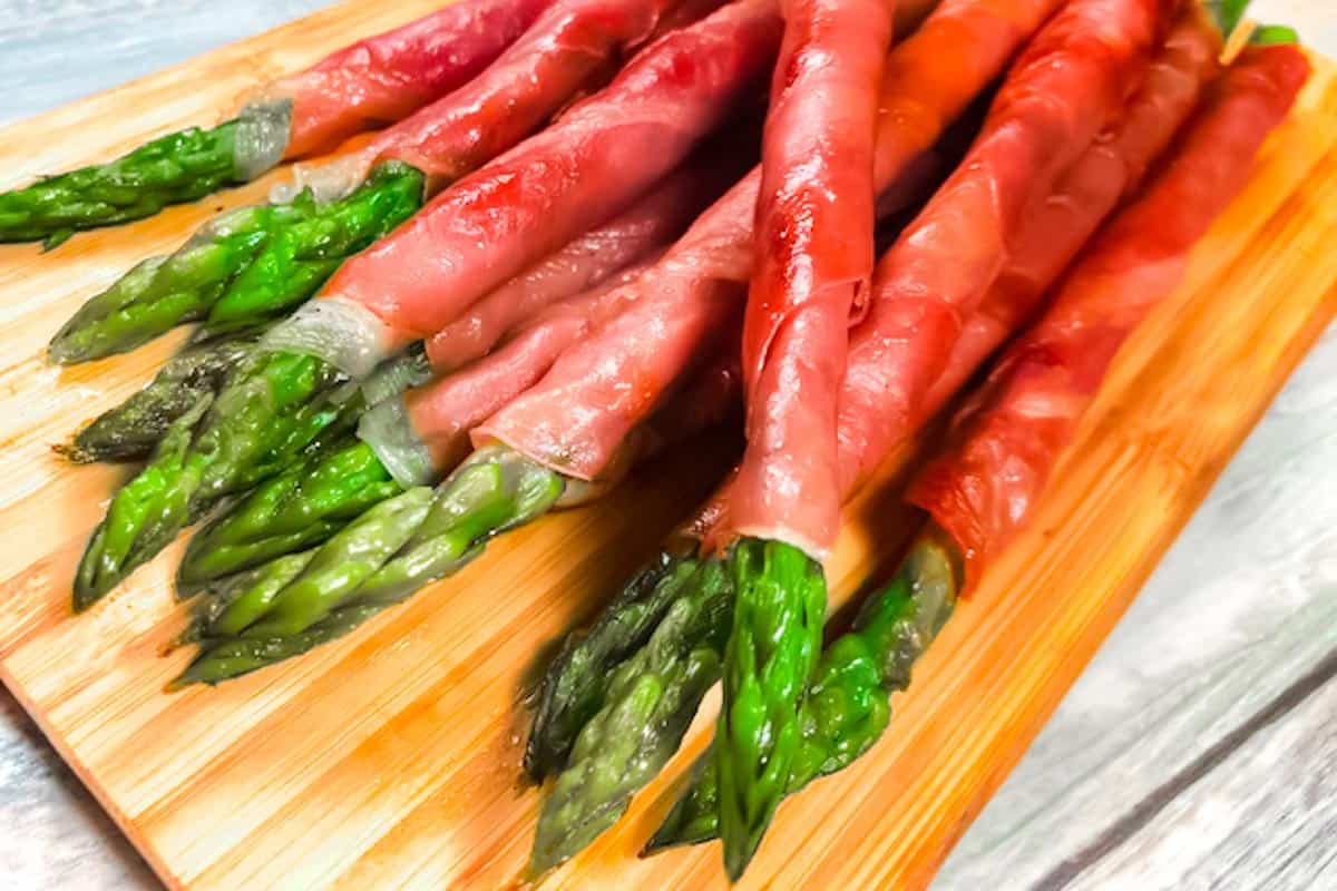 Closeup of a dozen crisped prosciutto wrapped asparagus on a bamboo cutting board.