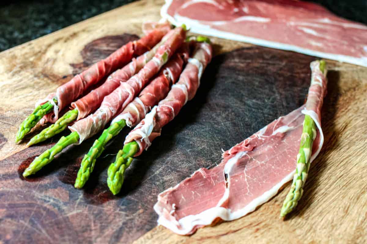 Blanched asparagus on a board, wrapped diagonally in prosciutto before crisping, with one stalk partially wrapped showing the process.