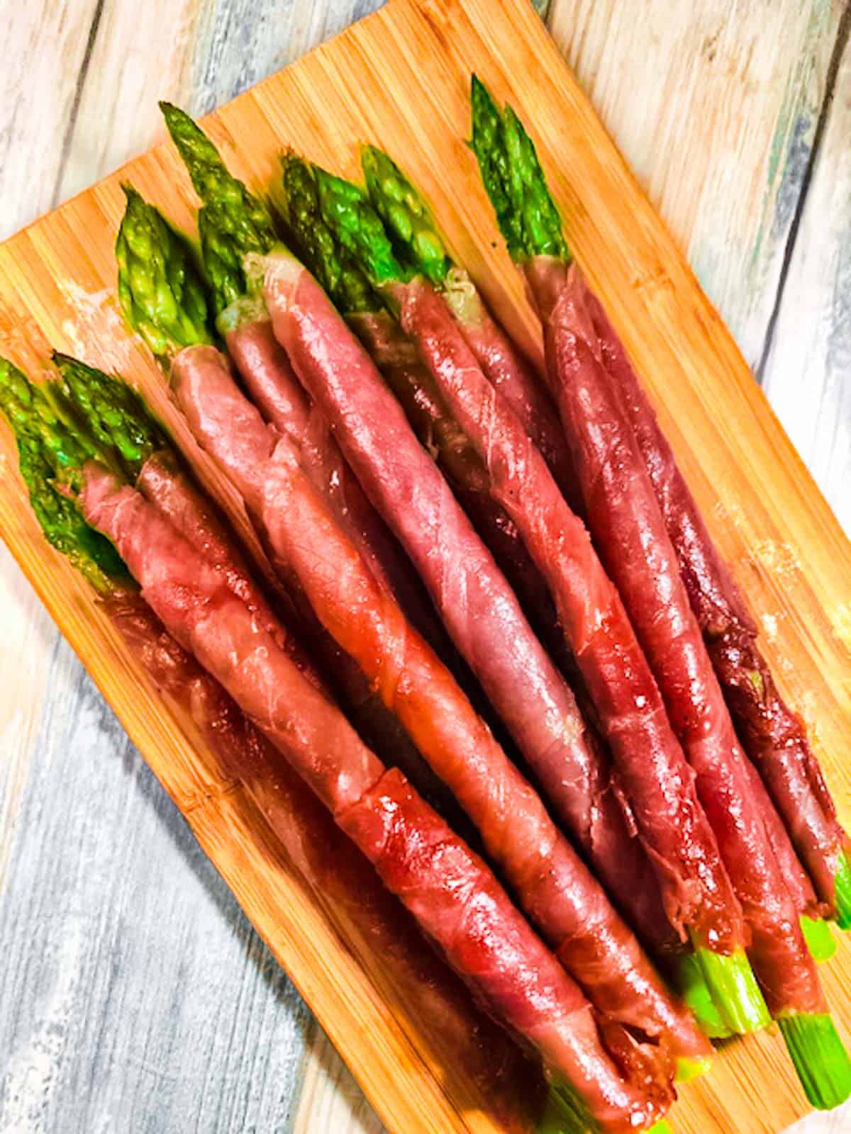 Overhead view of a dozen stalks of prosciutto wrapped asparagus sautéed crispy on the stovetop, and stacked on a cutting board.