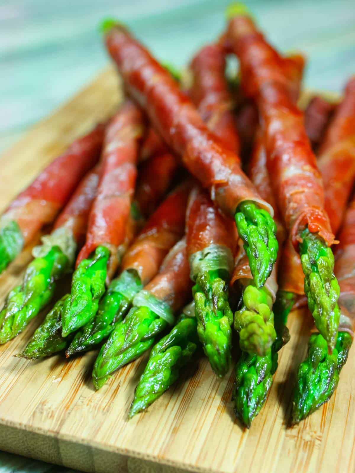 Asparagus wrapped in prosciutto stacked on a cutting board.