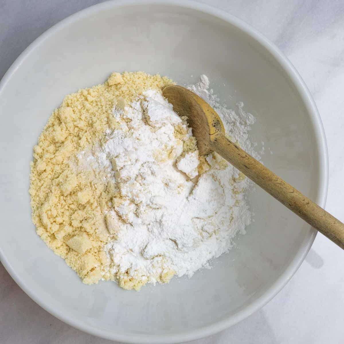 Process step showing dry ingredients for the cake in a mixing bowl with a wooden spoon in the bowl.