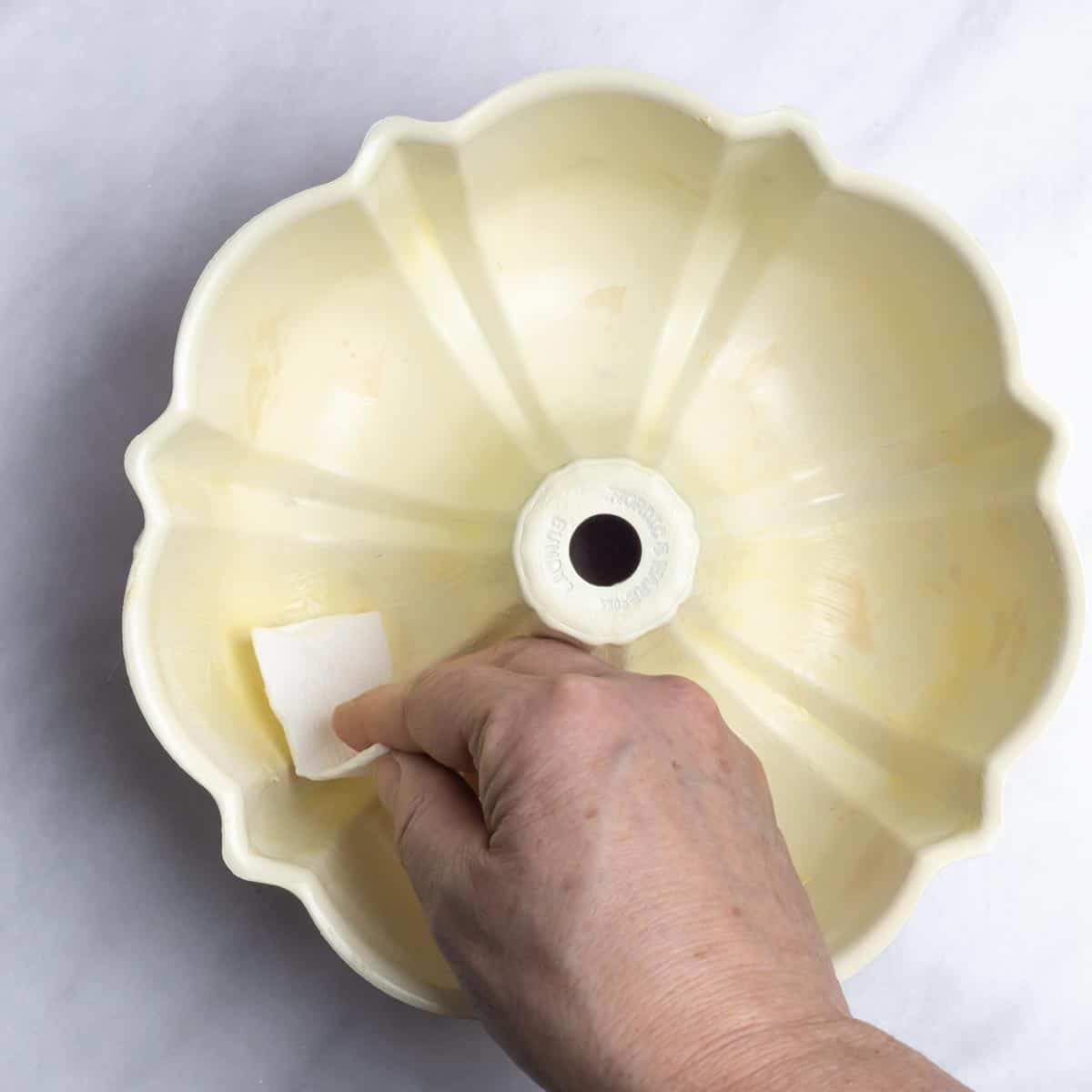 Hand with a paper towel piece greasing the inside of a bundt pan with butter.
