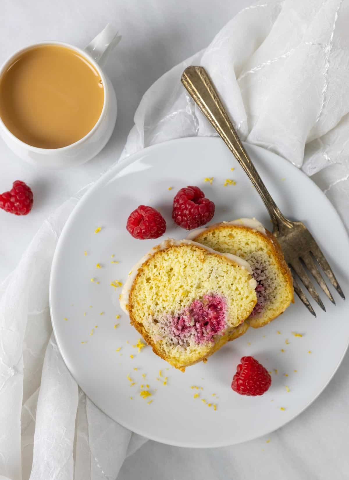 White plate with two slices of glazed keto bundt cake, a fork, and raspberries with lemon zest around the slices.