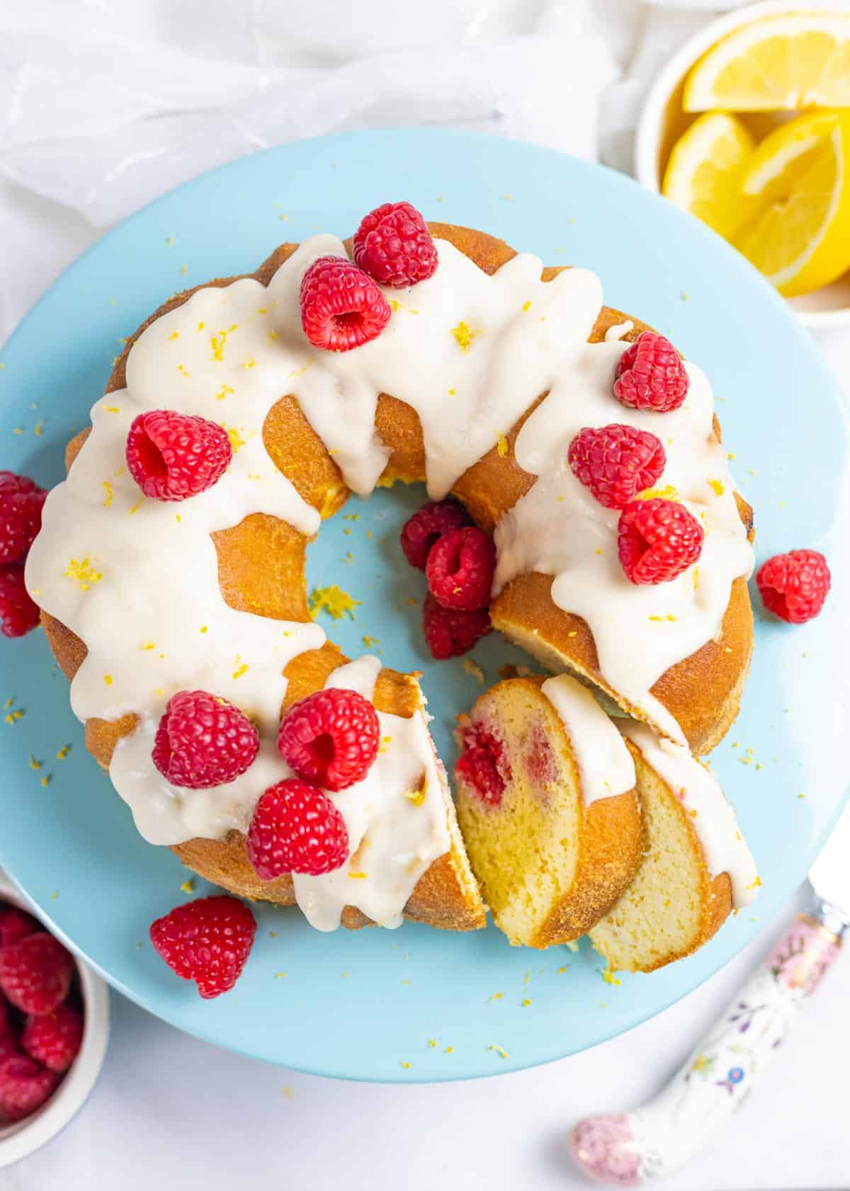 Lemon raspberry bundt cake on a blue cake pedestal topped with lemon glaze and whole raspberries.