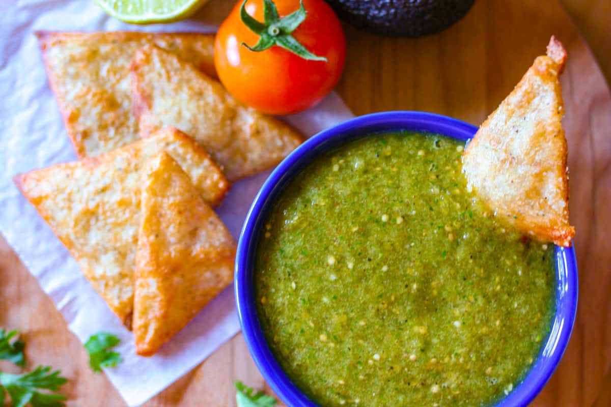 Tomatillo green chili salsa verde in a blue bowl with a side of keto chips, a tomato and an avocado.