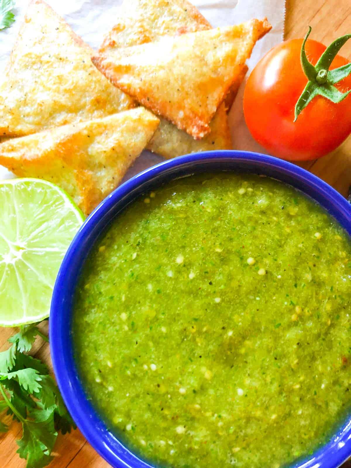 Salsa verde with tomatillos served in a blue bowl with a side of keto chips, with a whole tomato and half a lime next to the bowl.