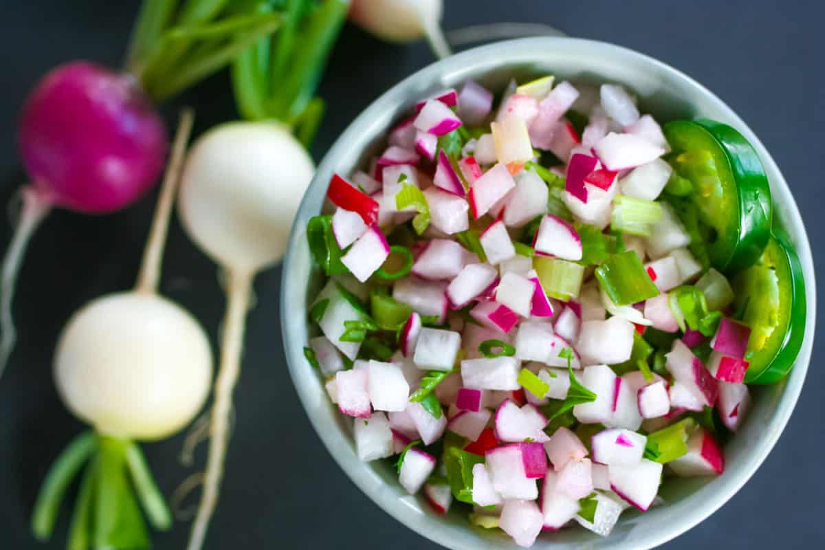 Blue bowl of radish salsa with sliced jalapeno garnish and whole white and red radishes on the side.
