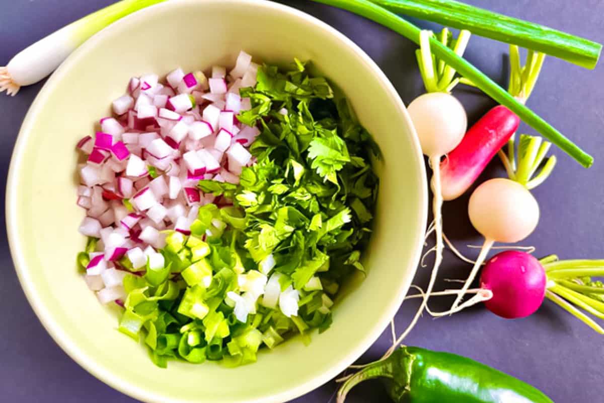 Ingredients for the salsa including chopped radish, green onion and cilantro in a yellow bowl with whole radishes and a jalapeno on the side.