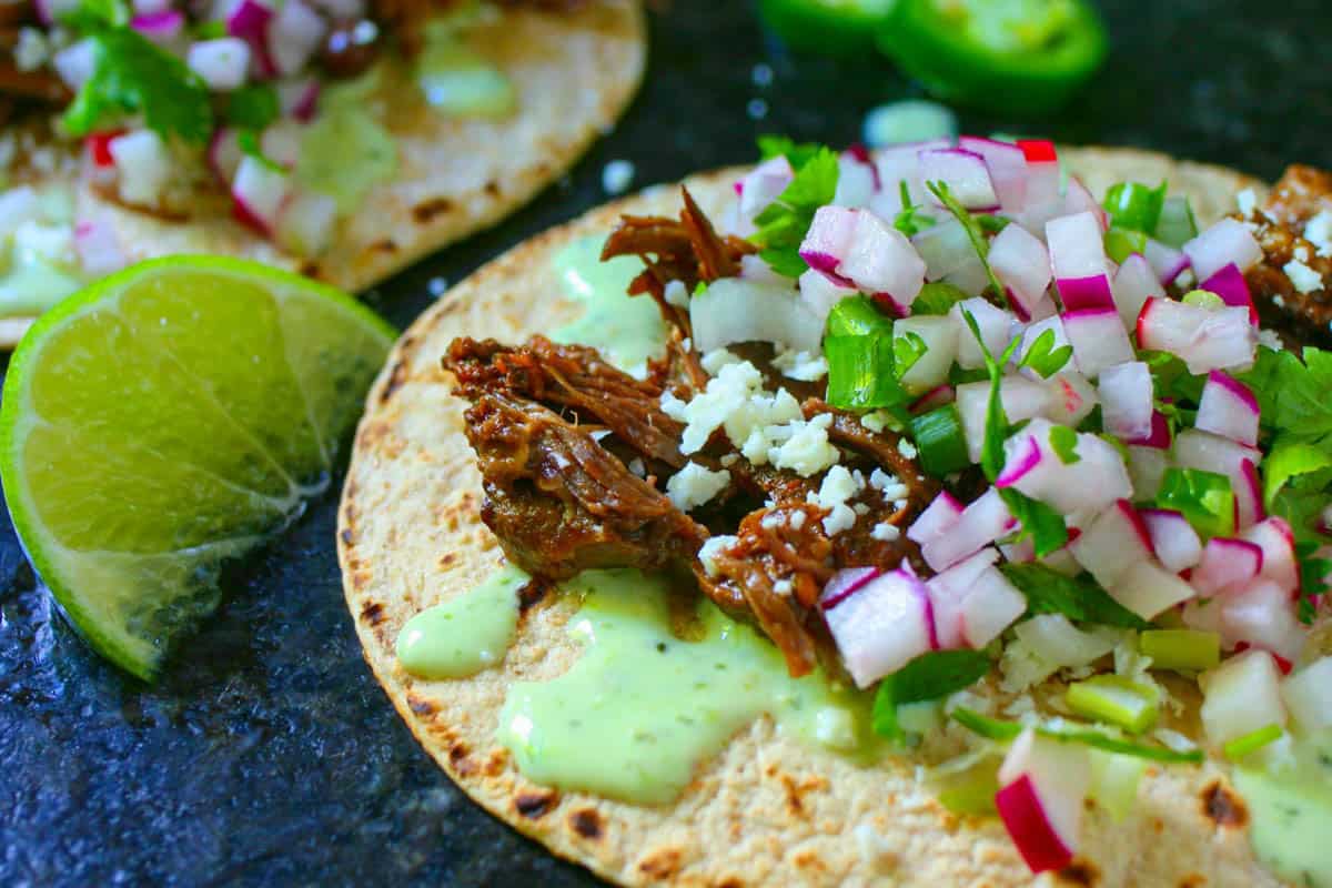 Flat beef taco tortillas with a lime wedge, crema dressing and radish salsa topped with cilantro on a black counter.