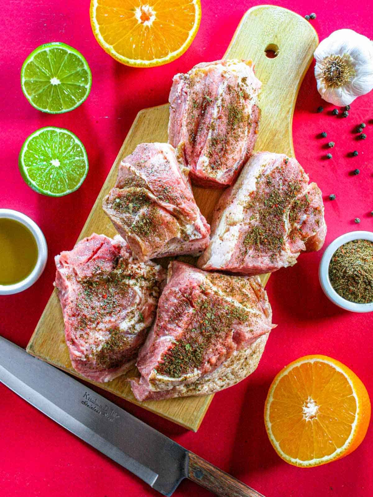 Large chunks of seasoned pork shoulder on a cutting board surrounded by small ramekins of olive oil and spice rub, halved orange and lime, a garlic head, with a chef's knife on a red background.