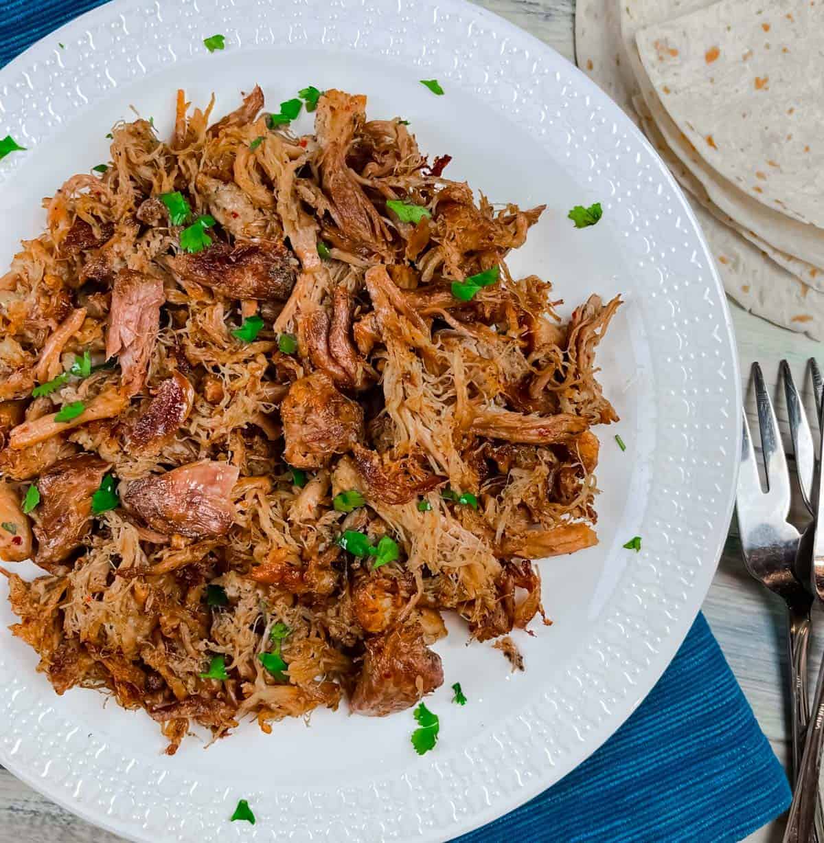 Crispy carnitas on a white plate with a side of tortillas and two serving forks.