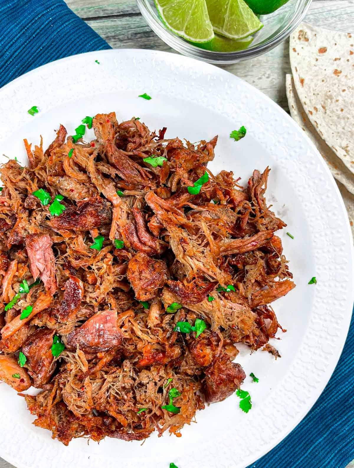 Shredded carnitas on a white plate with a sprinkle of fresh cilantro, a side of tortillas and glass bowl of lime wedges.
